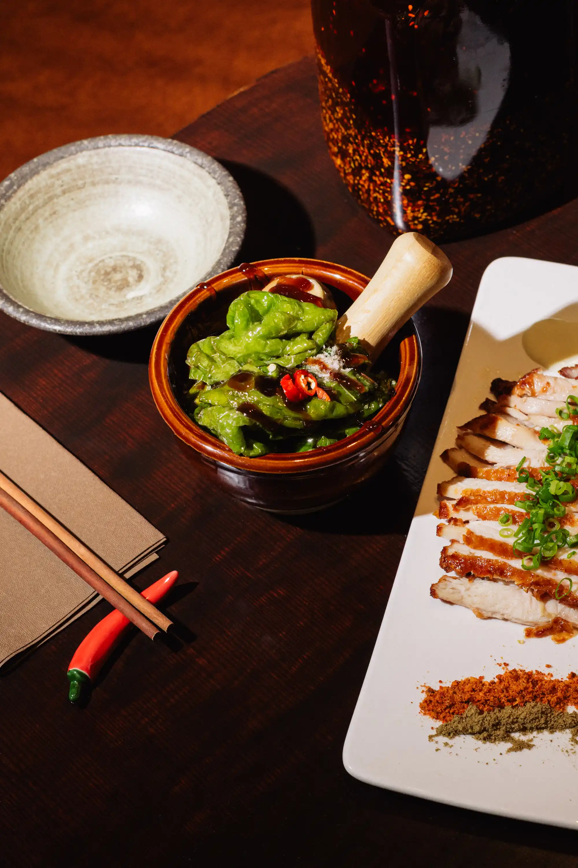 Sliced grilled chicken topped with chopped green onions and two spice powders on a white rectangular plate beside a ceramic bowl with lettuce and red chili garnished with sauce.