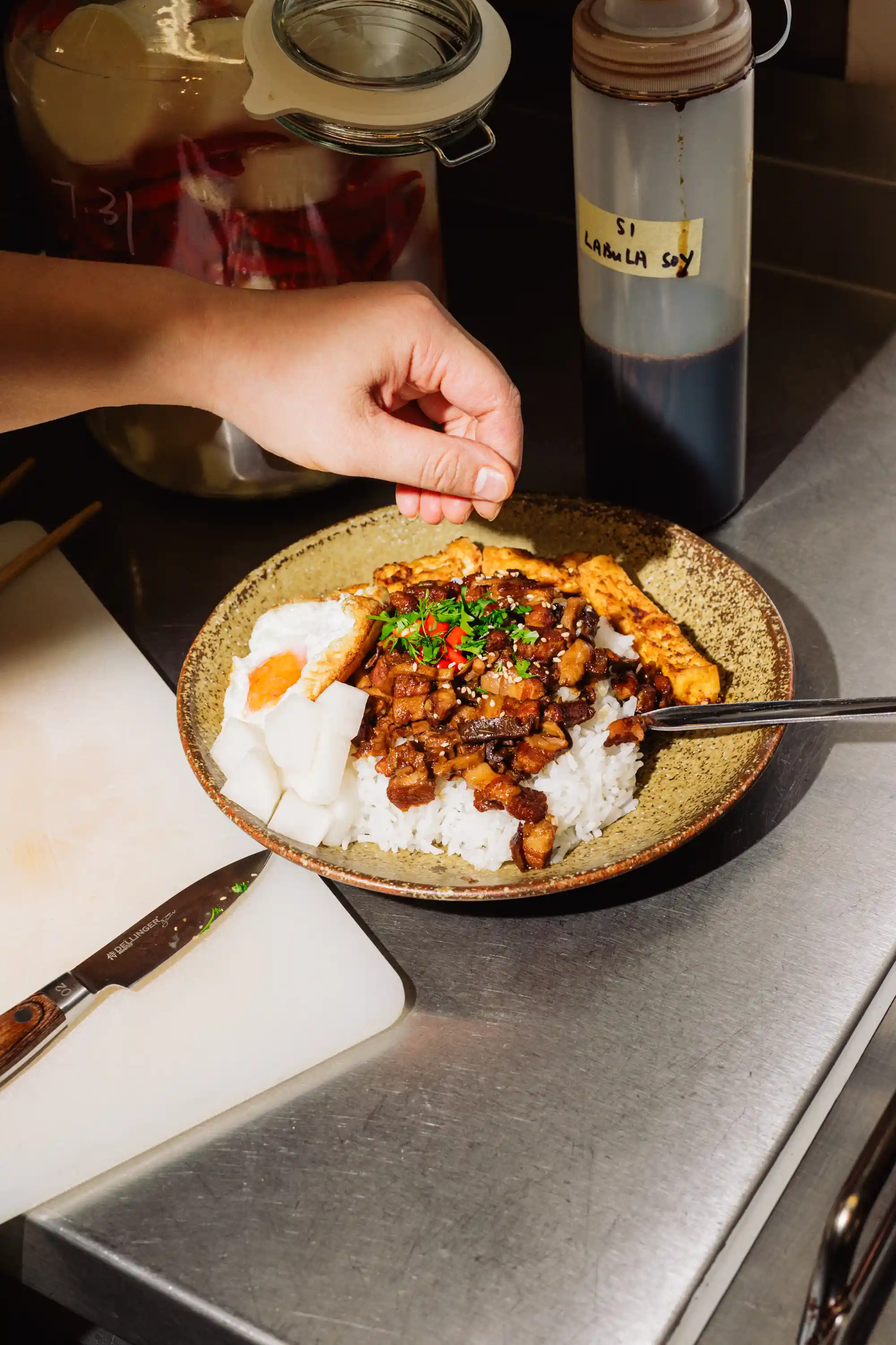 Hand sprinkling garnish over a bowl of rice topped with diced meat, a fried egg, tofu strips, and pickled radish on a stainless steel countertop.