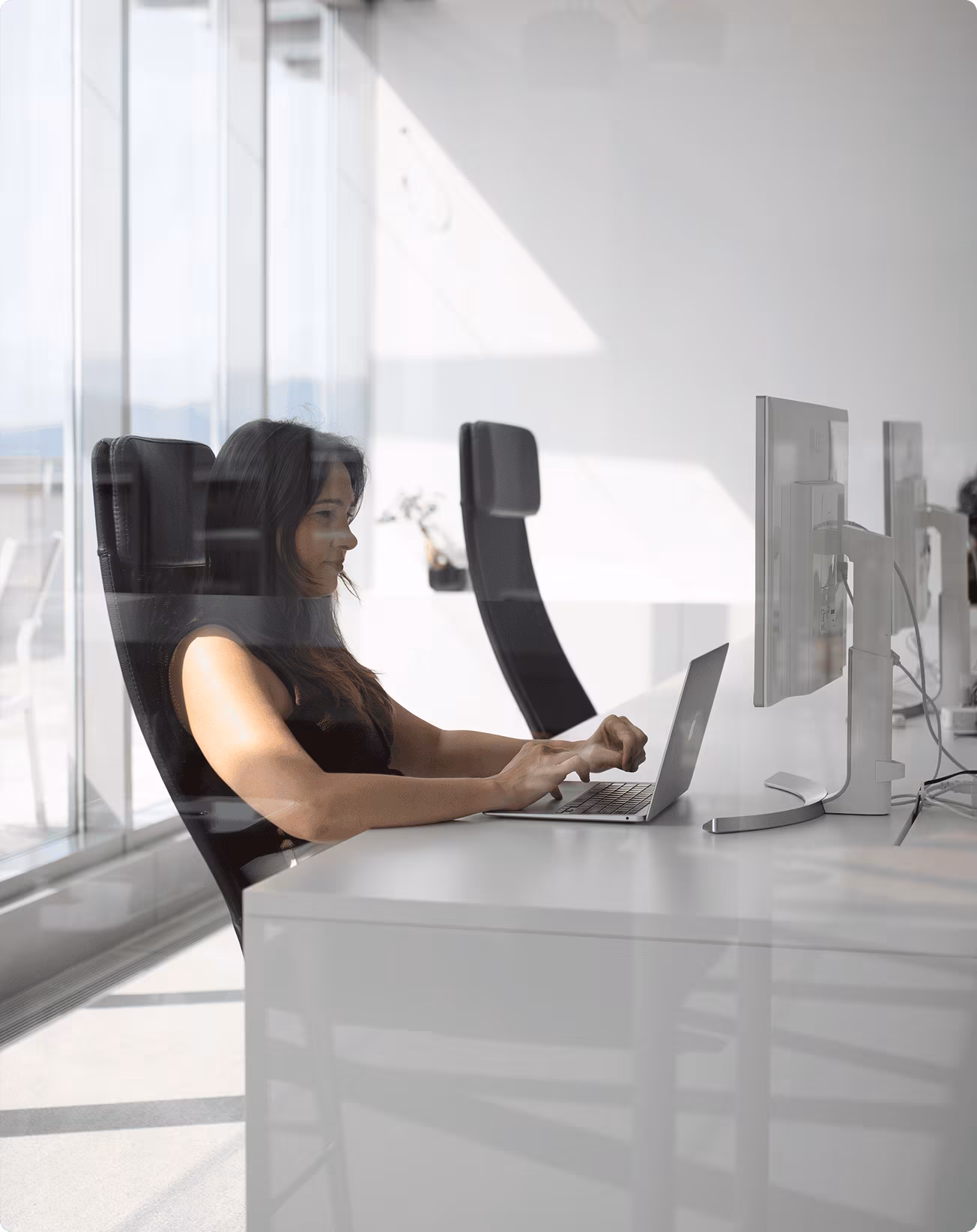 Woman working on a laptop at a white desk in a modern office with large windows and multiple computer monitors.