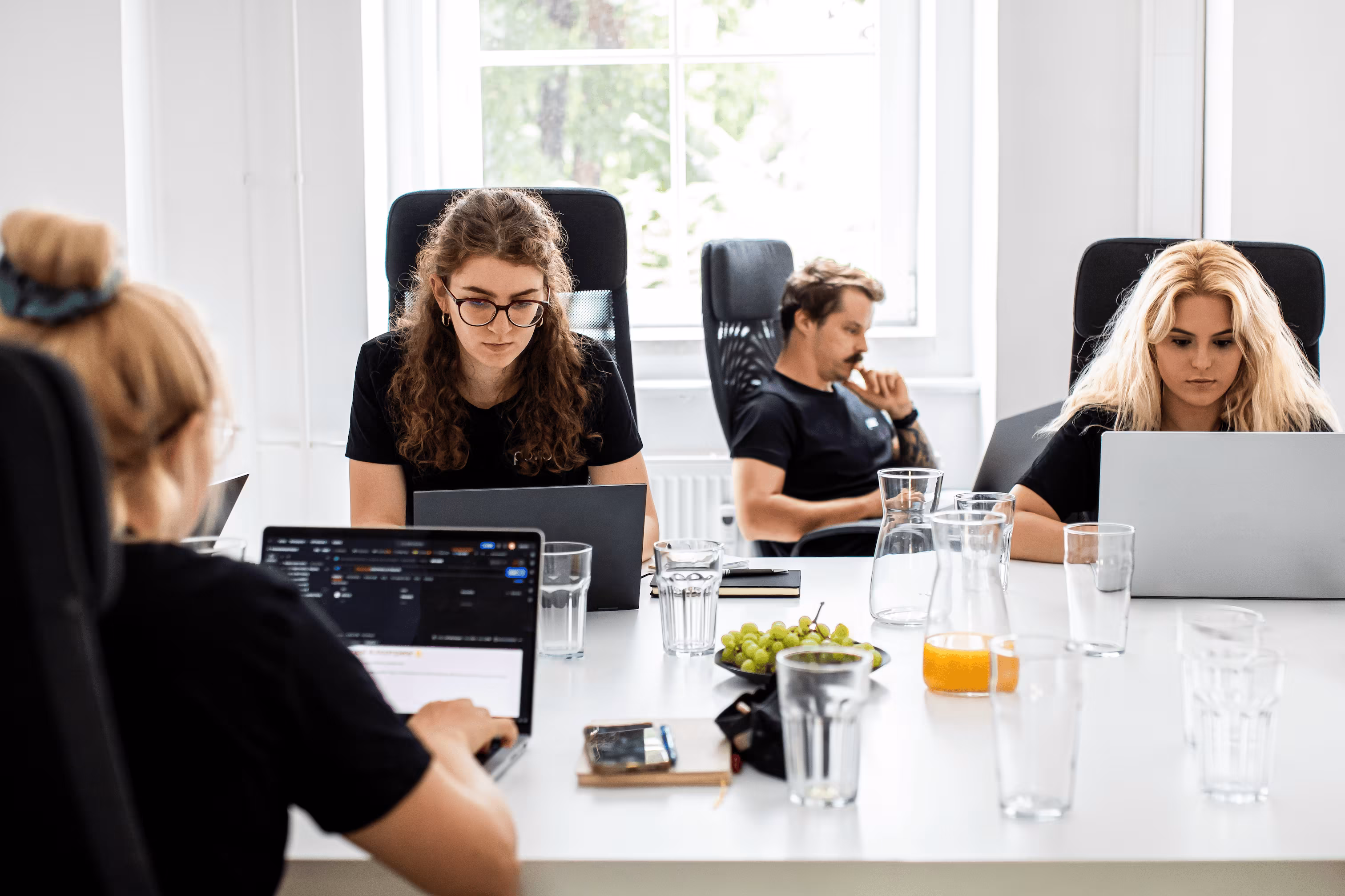 Four people focused on working with laptops around a white table in a bright office space.