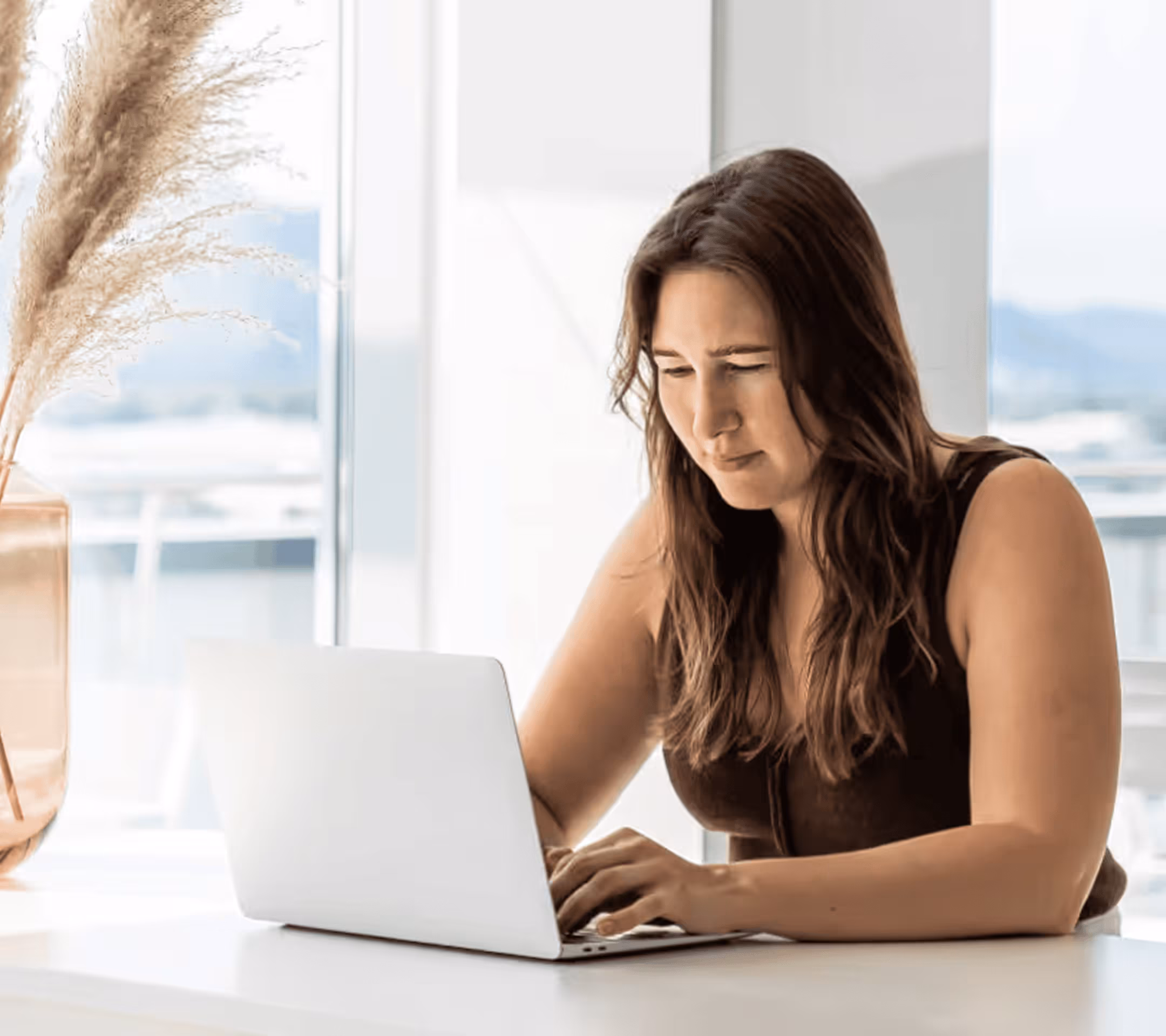 Woman with long brown hair working on a laptop at a white desk near a window with pampas grass in a vase.