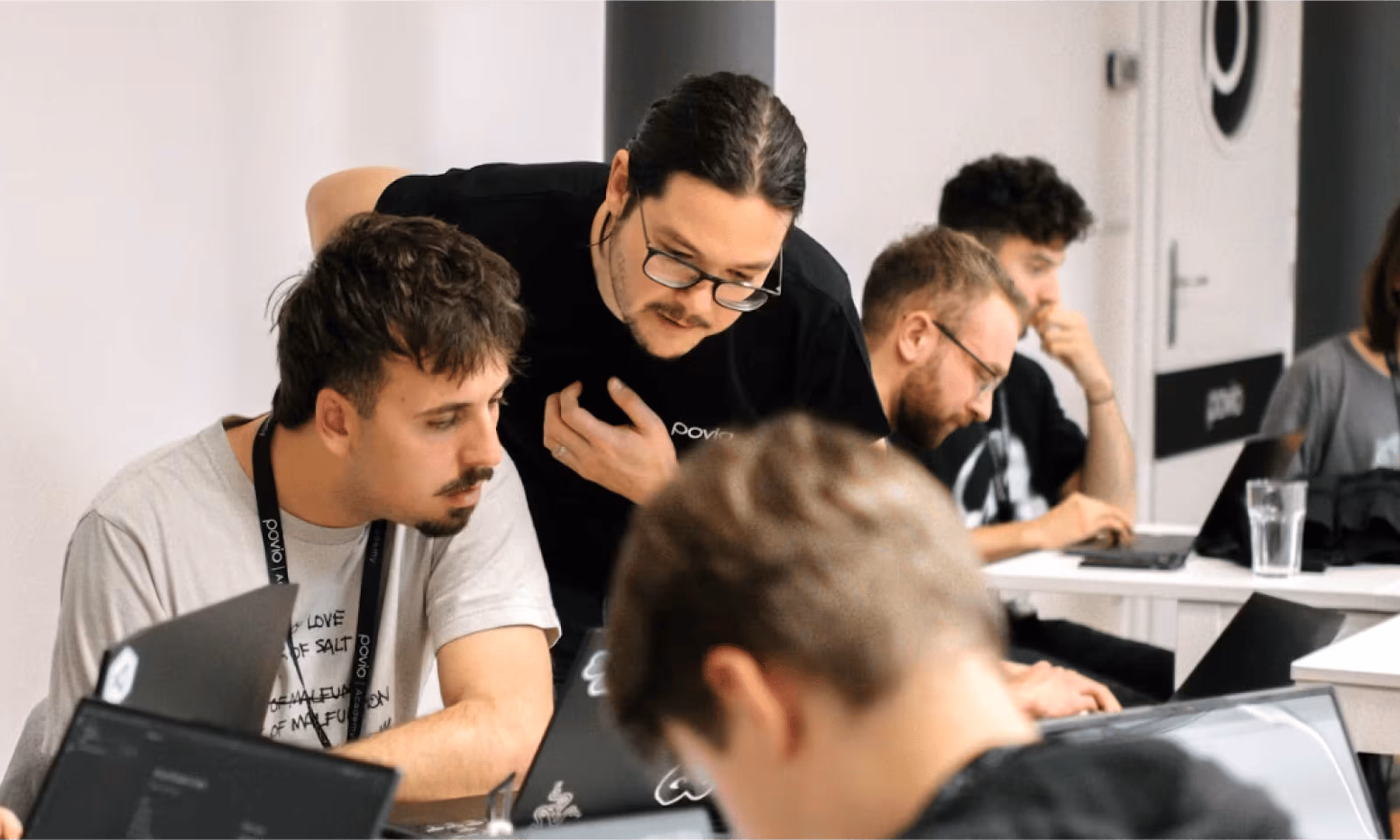 A man with glasses leans over to help another man working on a laptop in a collaborative office setting with other people using laptops in the background.