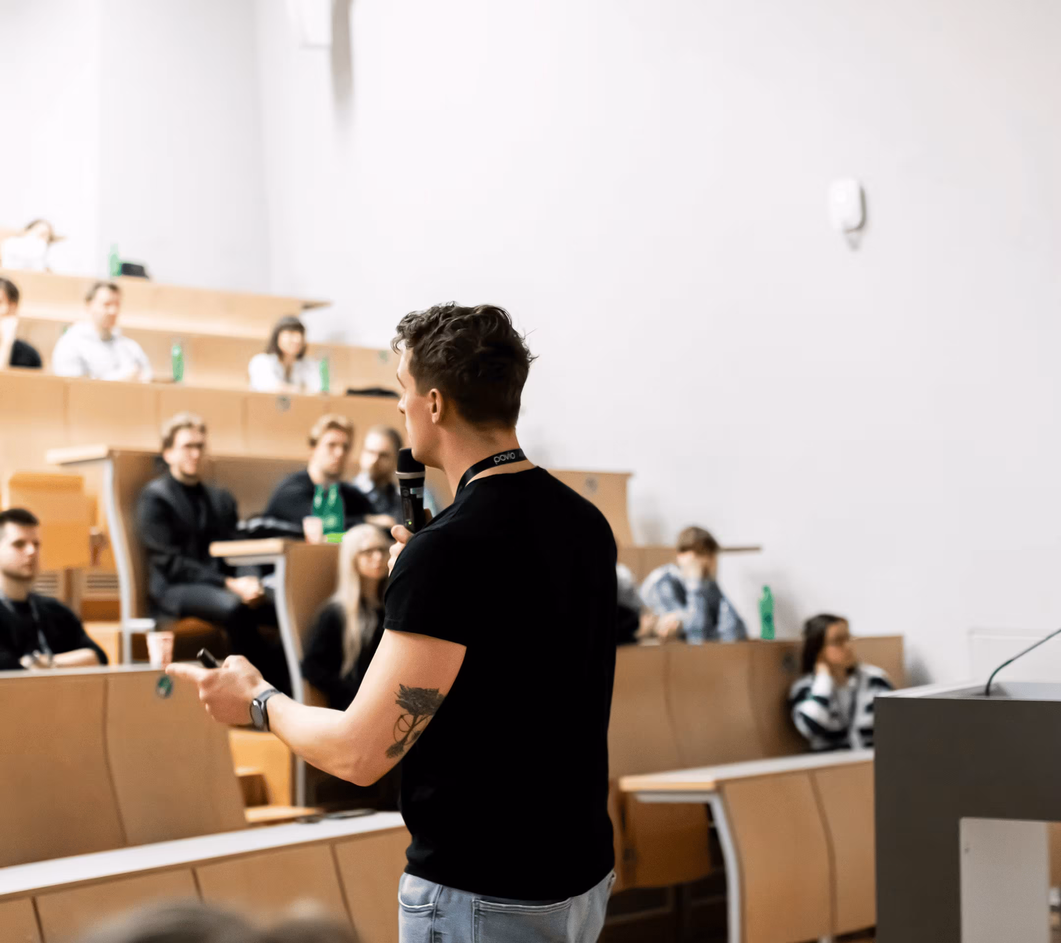 Man with a tattoo on his arm speaking into a microphone in a lecture hall with seated audience.