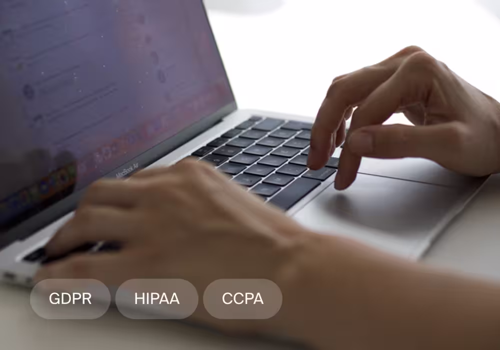Person typing on a MacBook Air laptop keyboard with GDPR, HIPAA, and CCPA tags overlay.