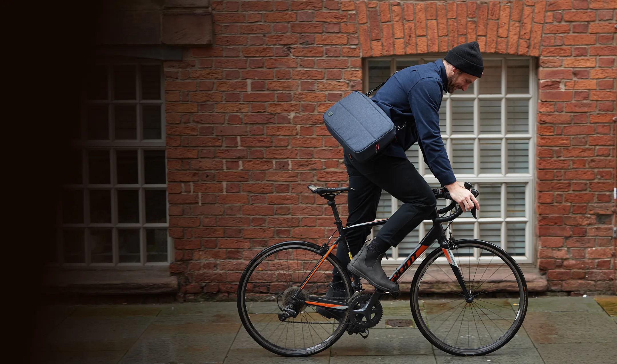 Man riding a black bike with a blue jacket and hat on. Sporting a Hotbox crossbody satchel.
