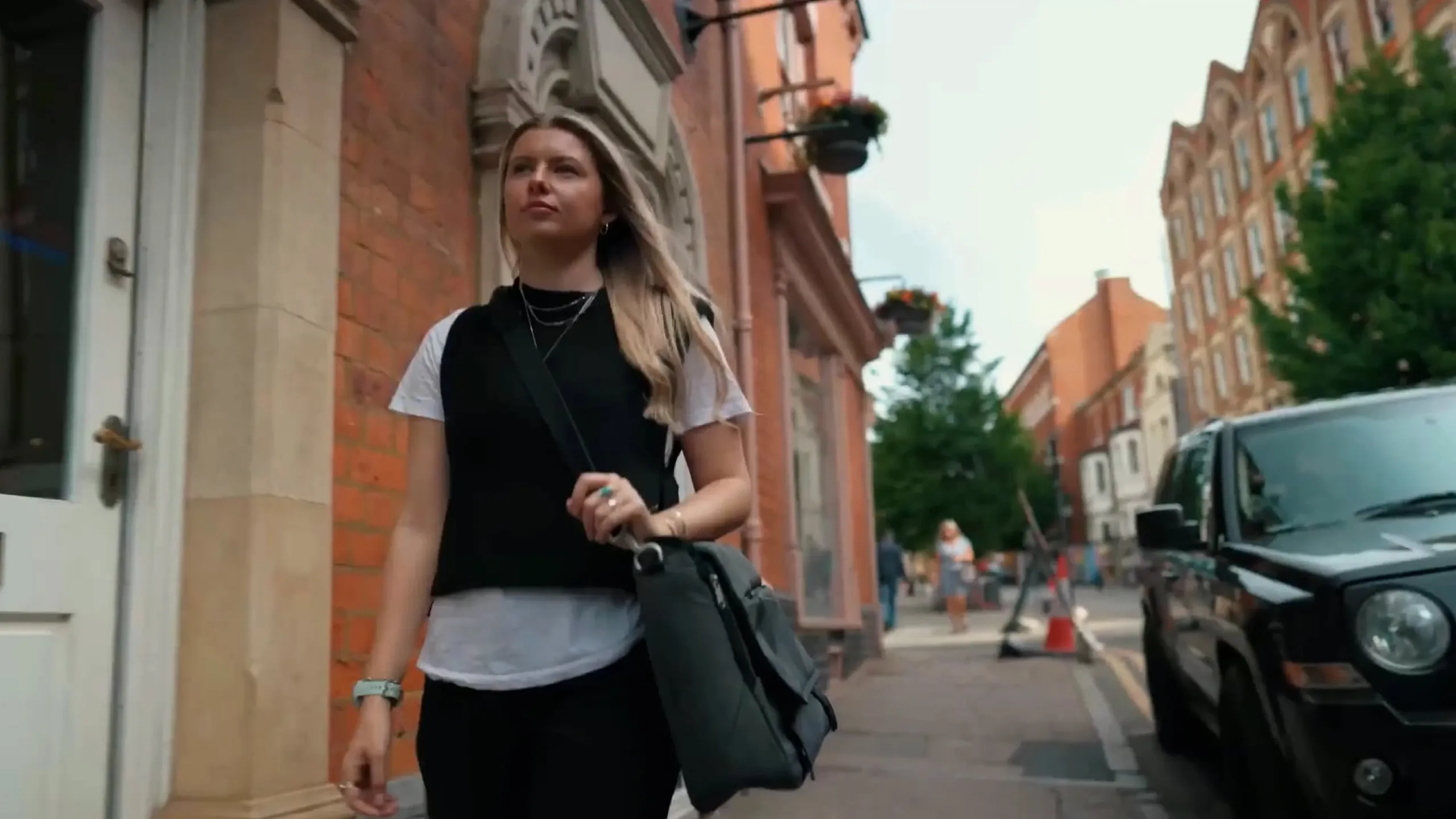 Young woman with long blonde hair walking on a city sidewalk beside brick buildings and a parked black vehicle.