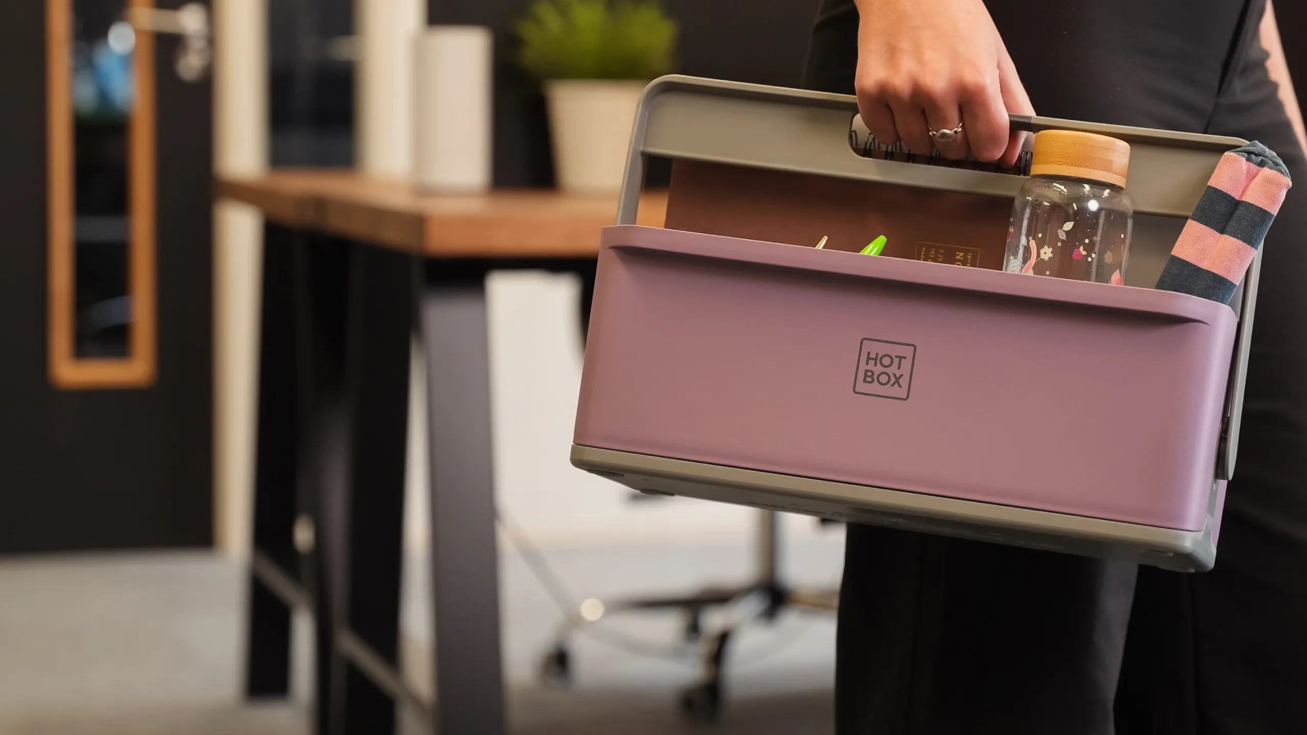 Person holding a purple Hotbox portable desk caddy filled with office supplies including a notebook, a water bottle, and a rolled fabric item.
