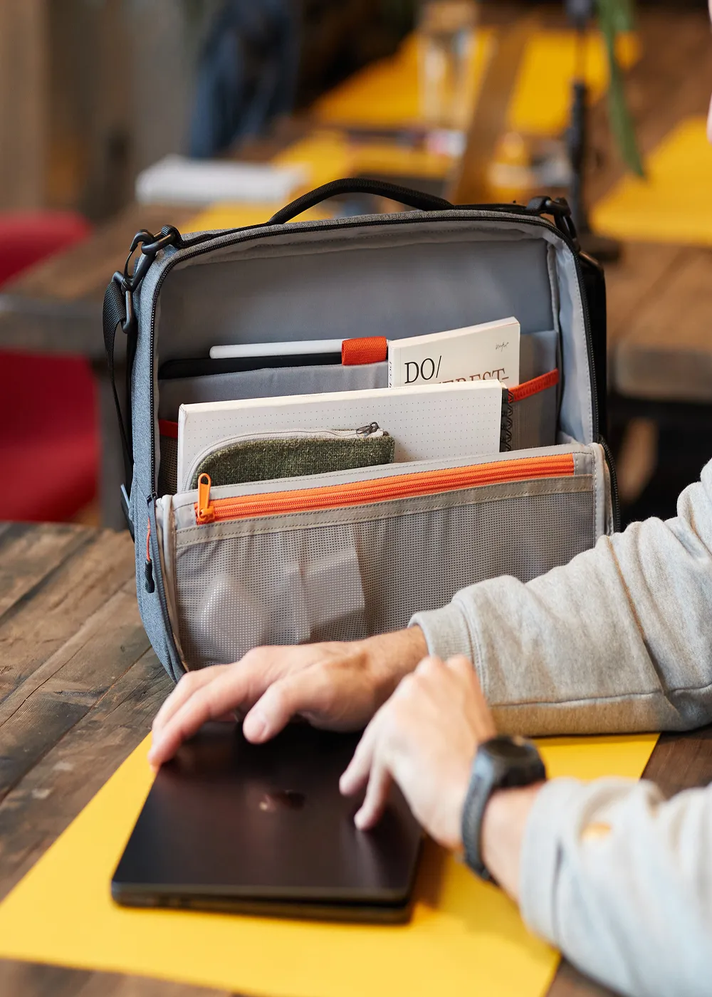 Open grey Adapt backpack on a table holding notebooks, a pencil, and a pencil case, with a person’s hands closing a black laptop nearby.