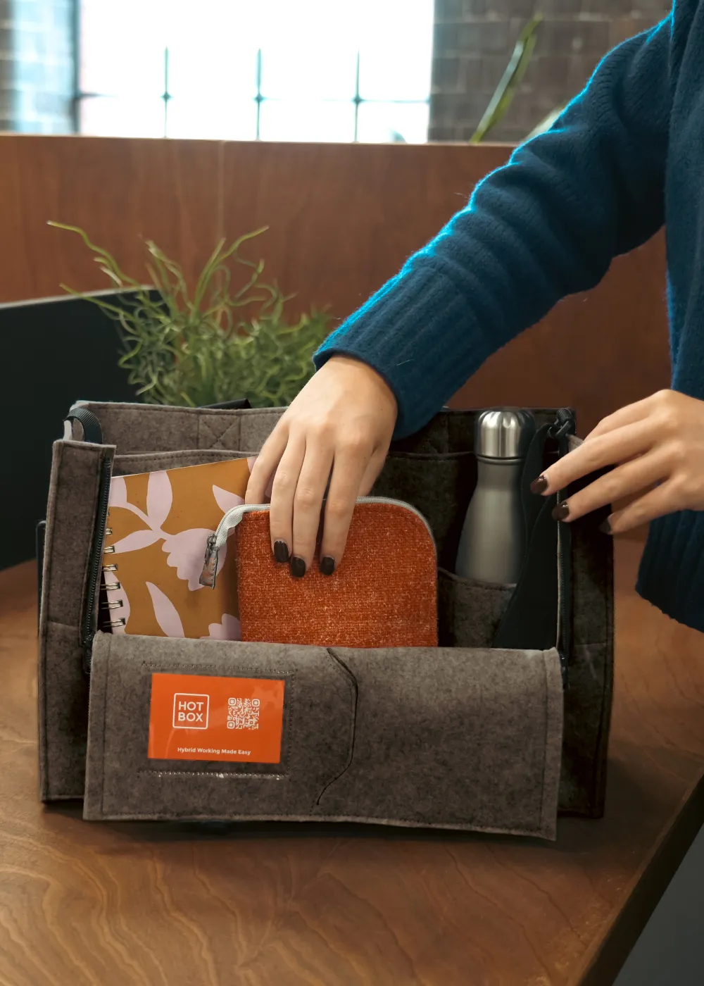 Person organising items including a notebook, pouch, and water bottle inside a grey Hotbox Flex caddy on a wooden table.