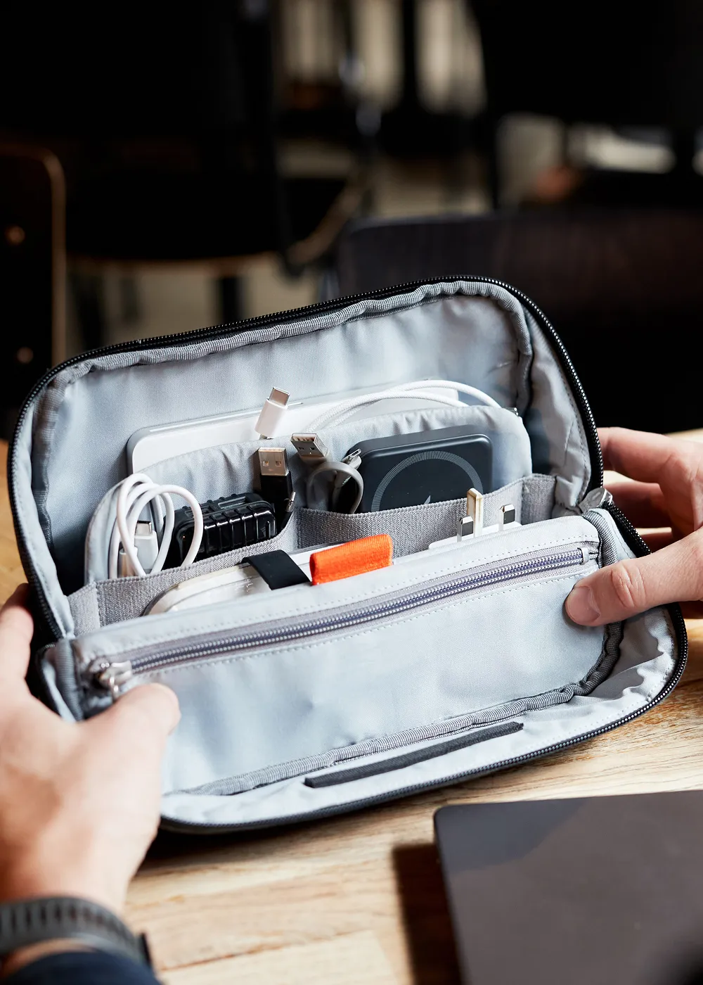 Hands holding an open grey Hotbox organiser pouch with various electronic cables, adapters, and a charging pad neatly stored inside.