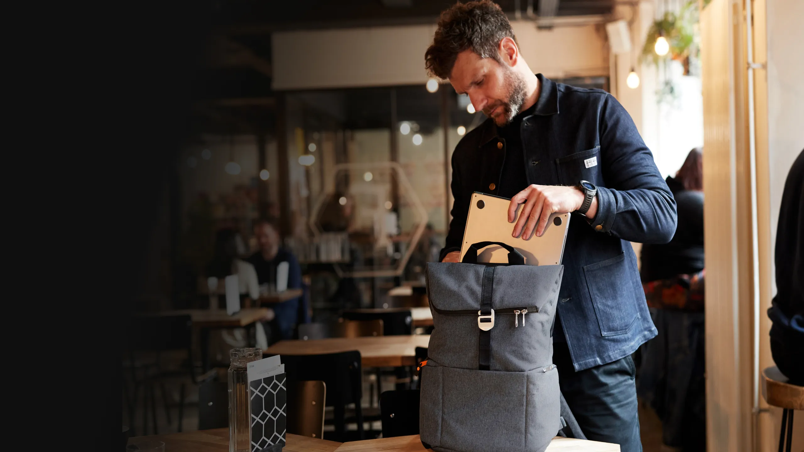 Man placing a laptop into a gray Shuttle backpack at a café with tables and people in the background.