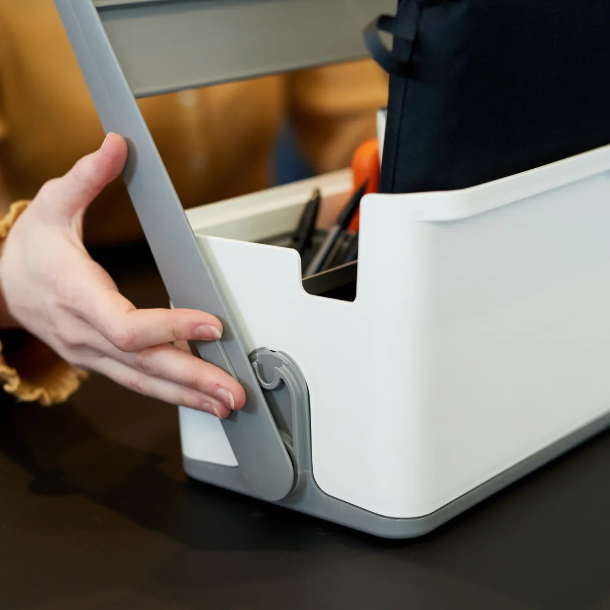 Hand adjusting the grey handle of a white plastic storage container holding pens and a black pouch.
