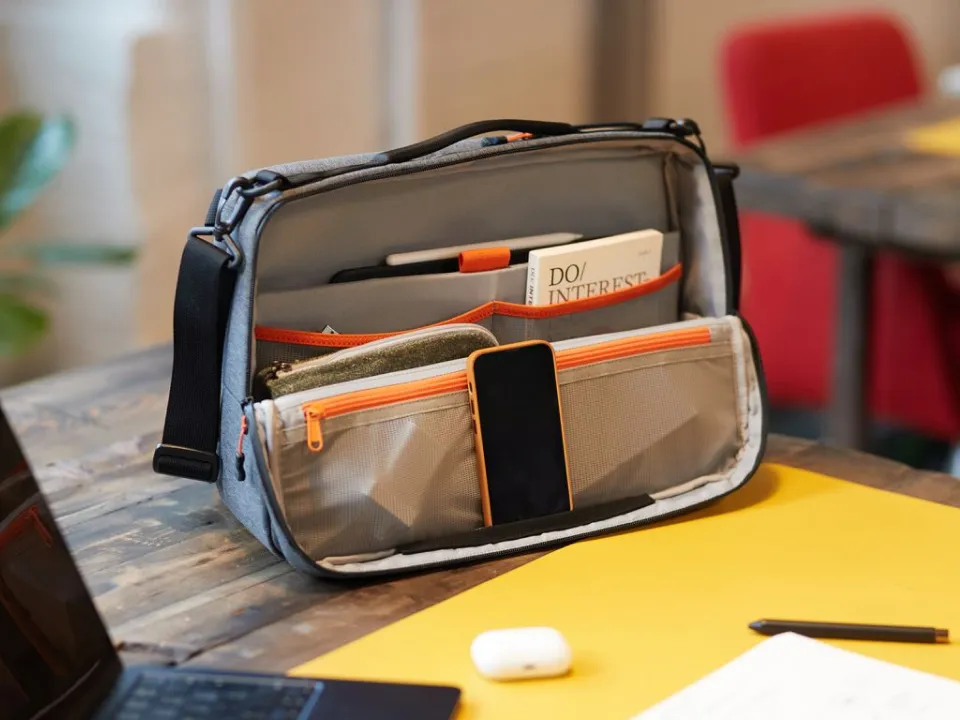 Opened grey shoulder bag with orange accents on a wooden desk, containing a phone, a book, a pen, and a pouch.
