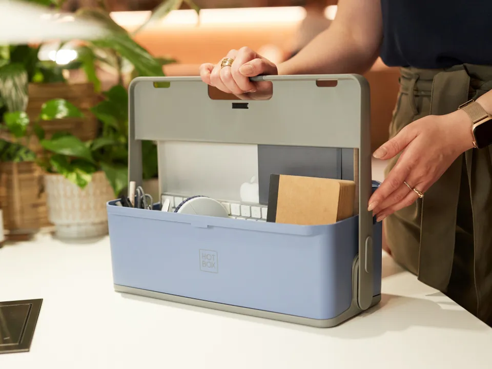 Person holding a blue Hotbox carrying case filled with office supplies, including a laptop, notebook, and keyboard.