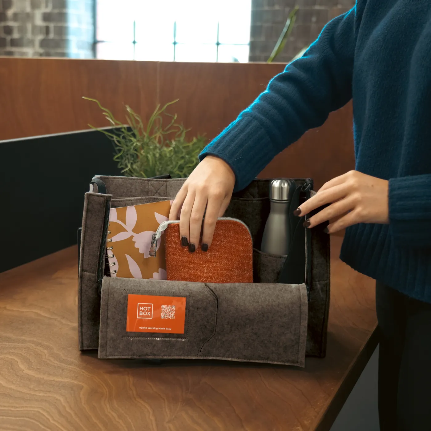 Person organising items including a notebook, pouch, and water bottle inside a grey Hotbox Flex caddy on a wooden table.