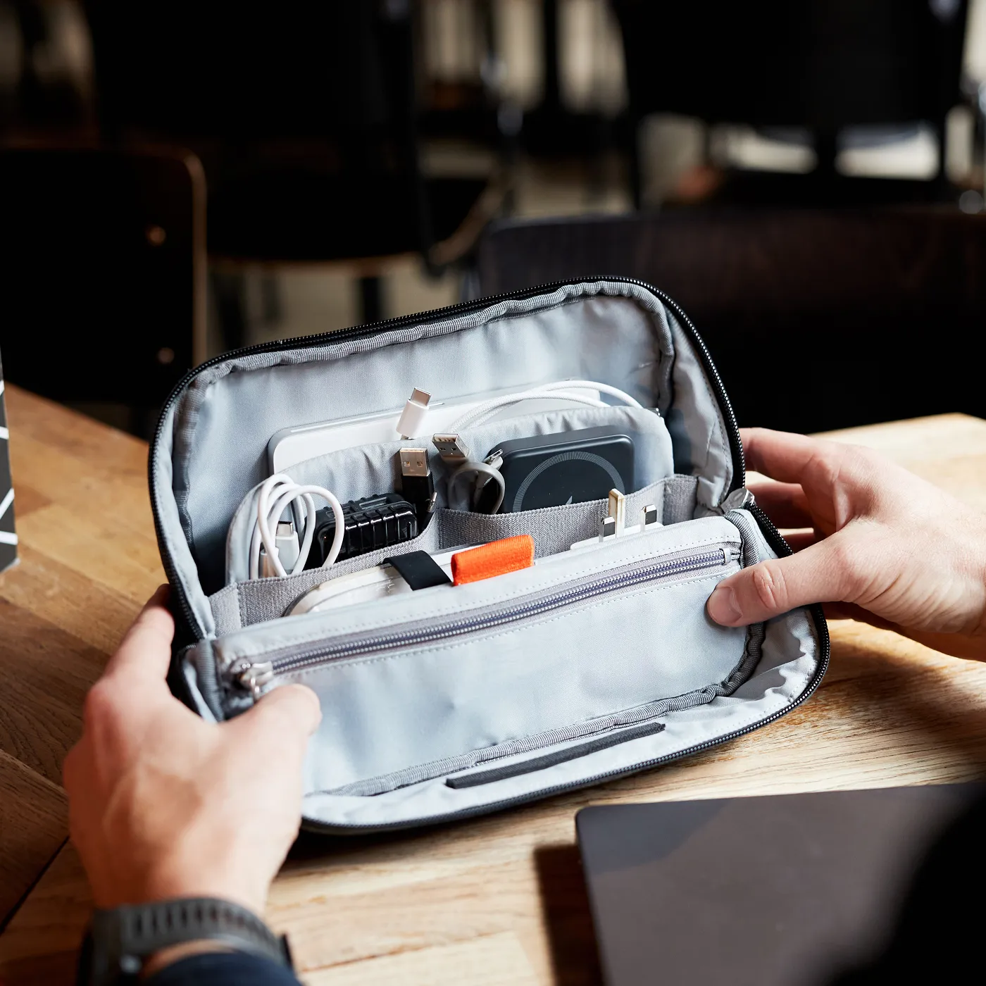 Hands holding an open grey Hotbox organiser pouch with various electronic cables, adapters, and a charging pad neatly stored inside.