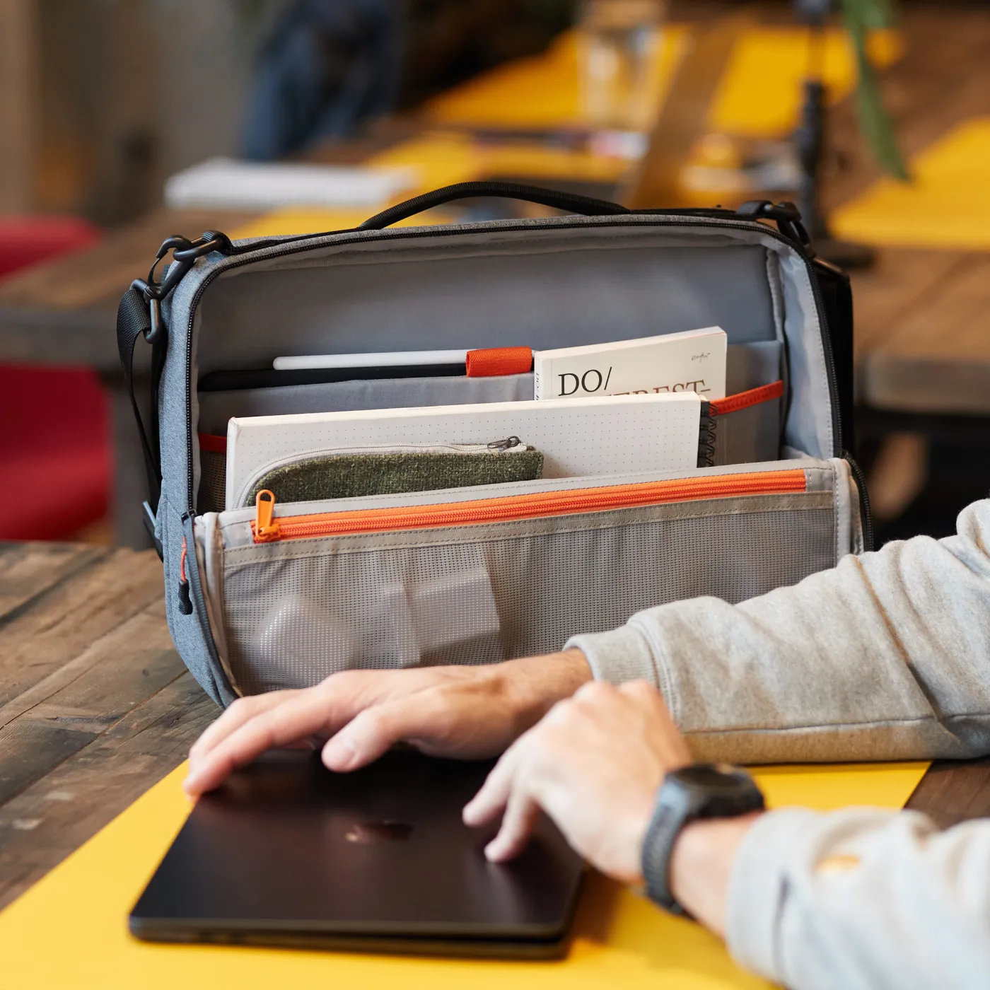 Open grey Adapt backpack on a table holding notebooks, a pencil, and a pencil case, with a person’s hands closing a black laptop nearby.