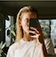 Person with light hair in a patterned shirt taking a selfie in a room with plants and shelves.