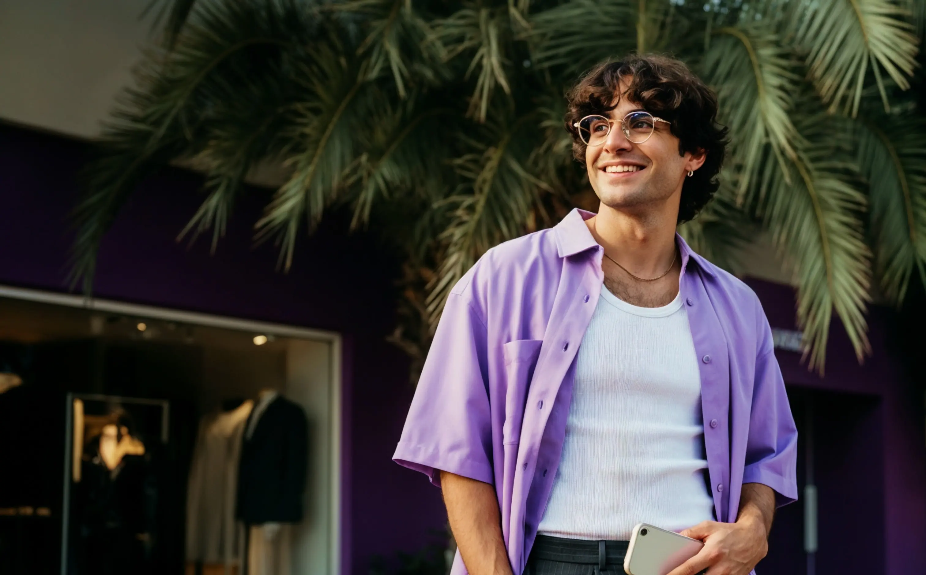 Smiling young man with curly hair wearing glasses, a purple shirt over a white tank top, standing in front of green palm leaves.