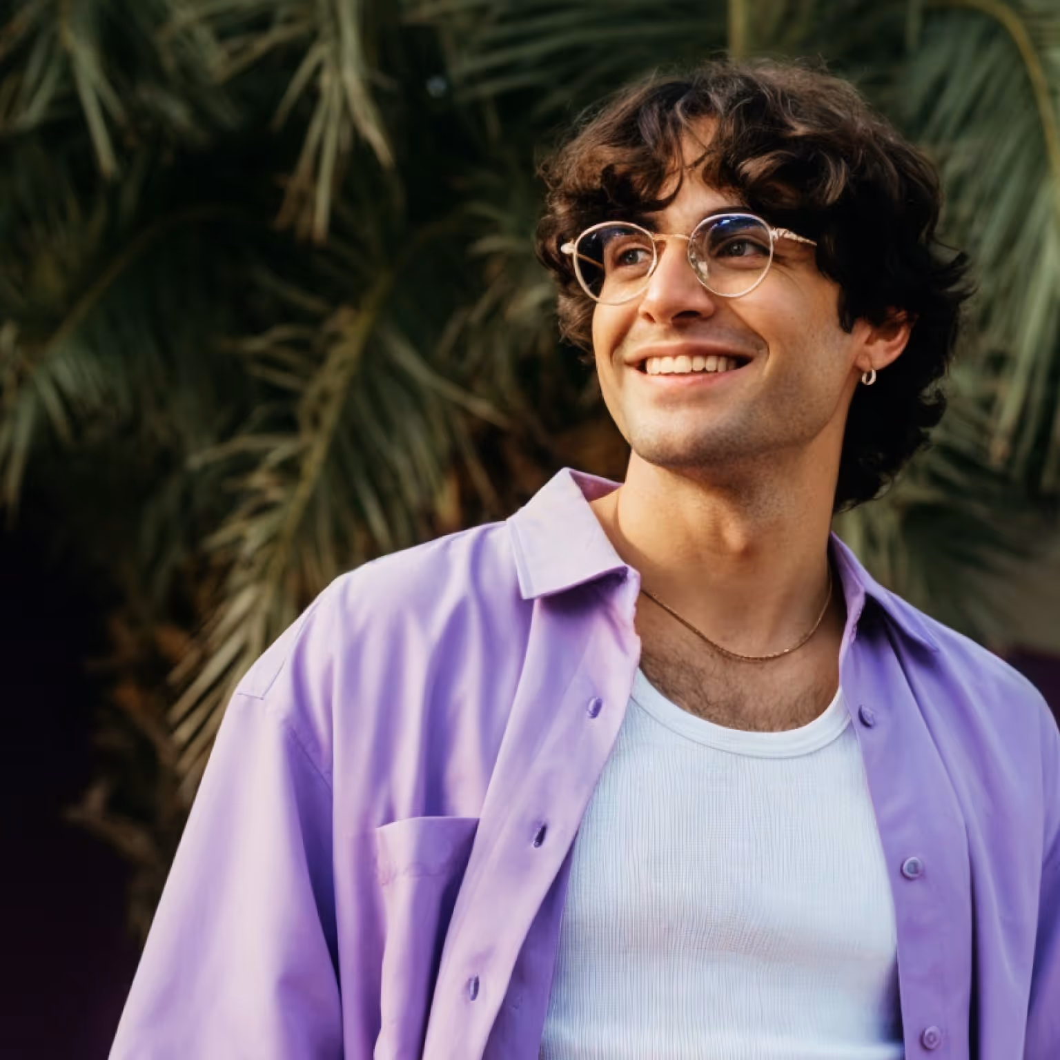 Smiling young man with curly hair wearing glasses, a purple shirt over a white tank top, standing in front of green palm leaves.