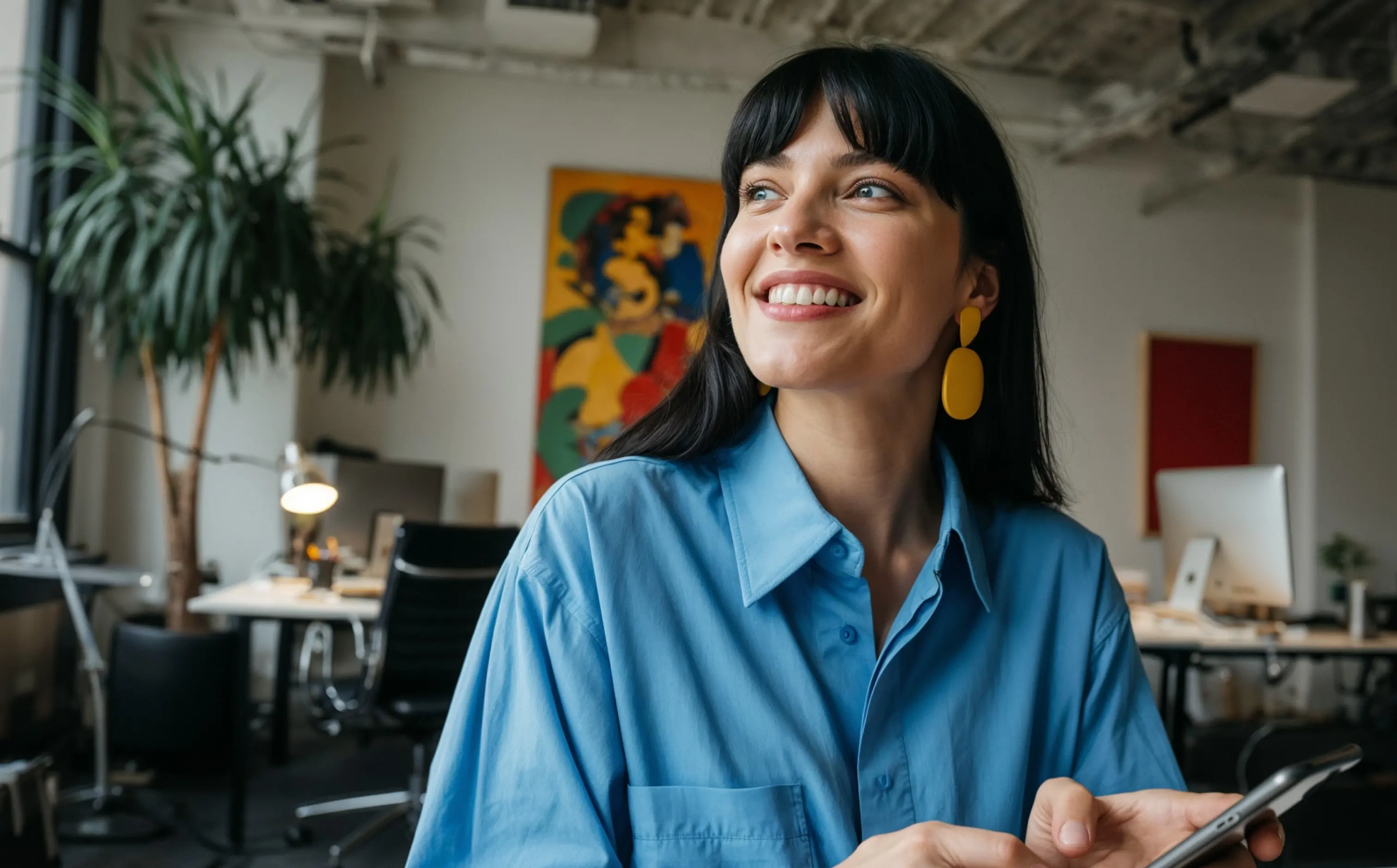 Smiling woman with black hair and yellow earrings holding a smartphone in a modern office.