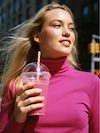 Smiling blonde woman in pink turtleneck holding a drink with a straw outdoors.