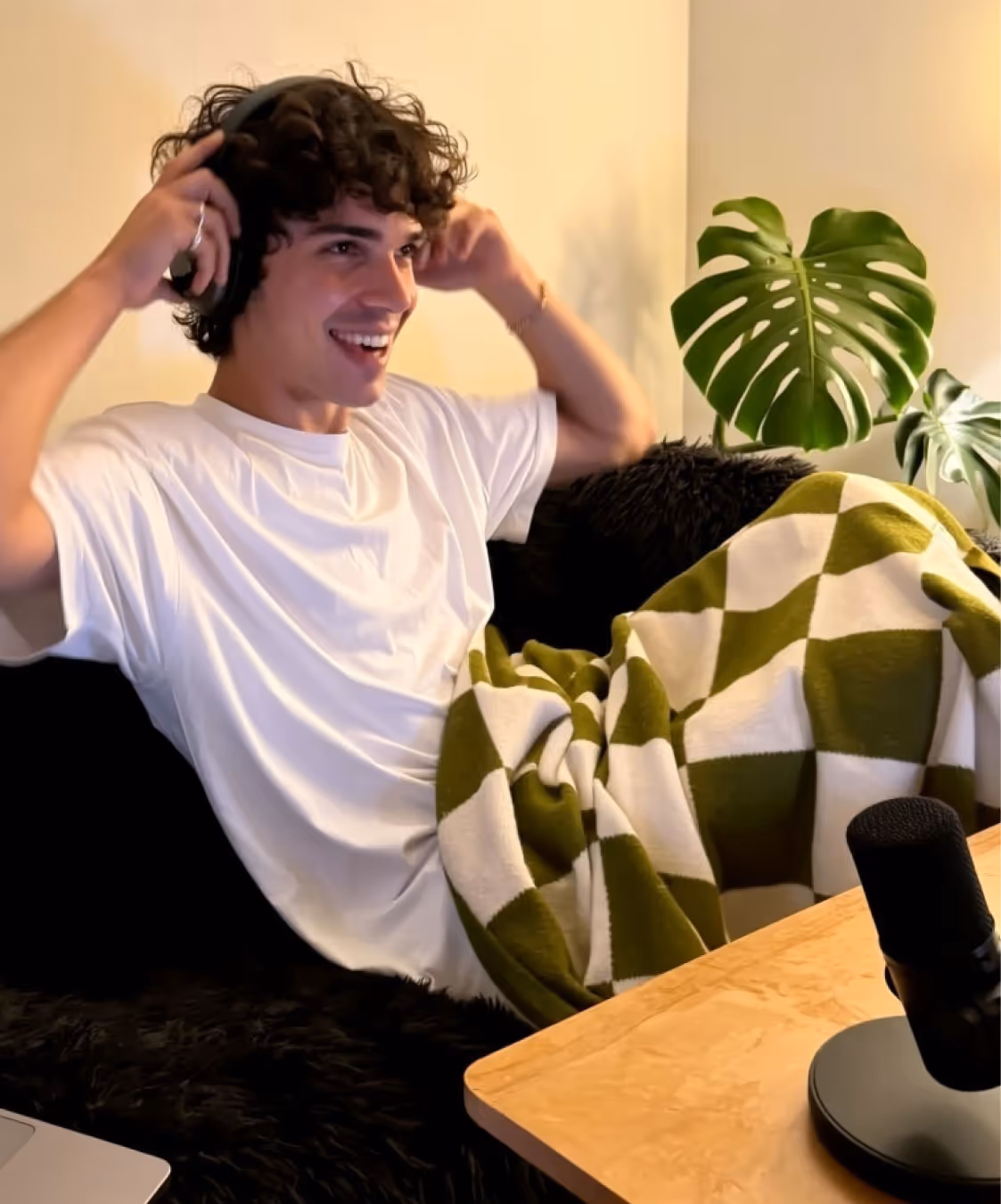 Young man with curly hair smiling and putting on headphones while sitting on a black couch with a green and white checkered blanket and a microphone on a nearby wooden table.