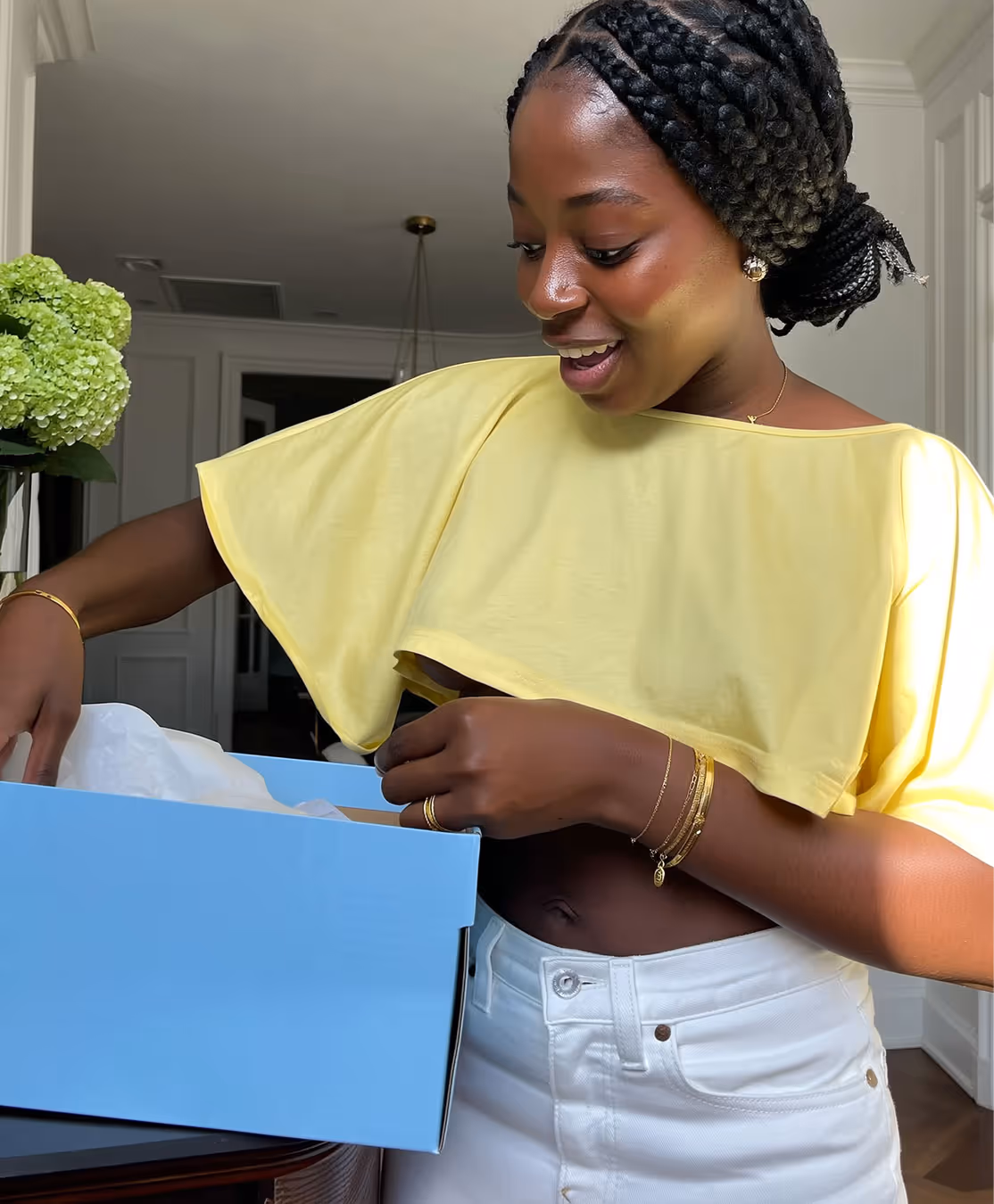 Smiling woman in yellow top and white pants looking into a light blue box indoors.