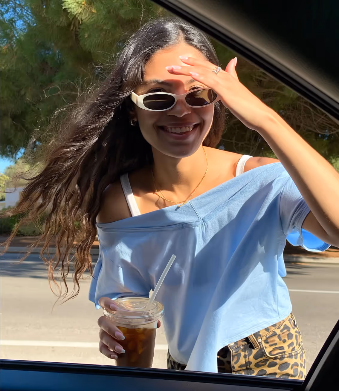 Smiling woman with long hair wearing white sunglasses and a blue off-shoulder top holding an iced drink outside a car window.