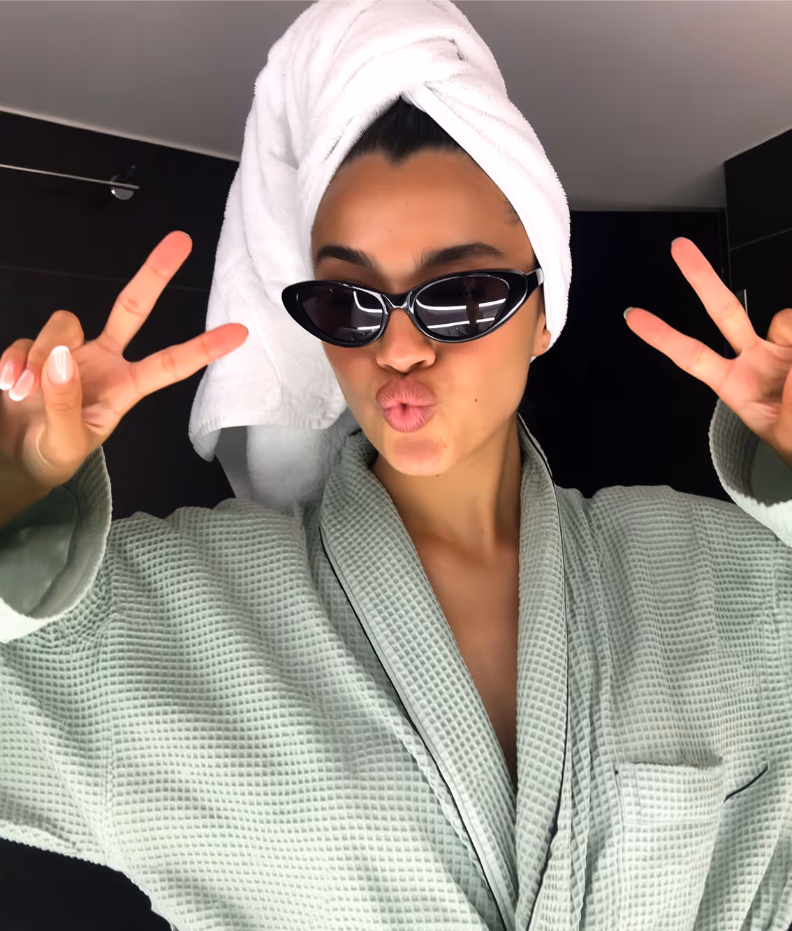 Woman wearing a light green robe, a white towel wrapped on her head, black cat-eye sunglasses, and making peace signs with both hands while puckering her lips.