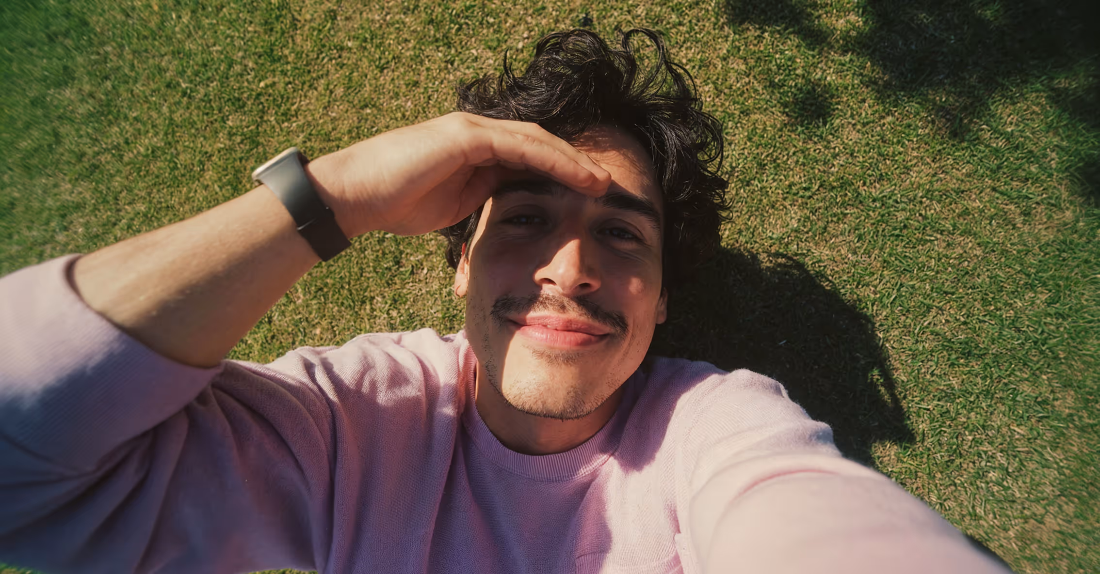 Young man lying on grass, shading his eyes from the sun with one hand and smiling at the camera.