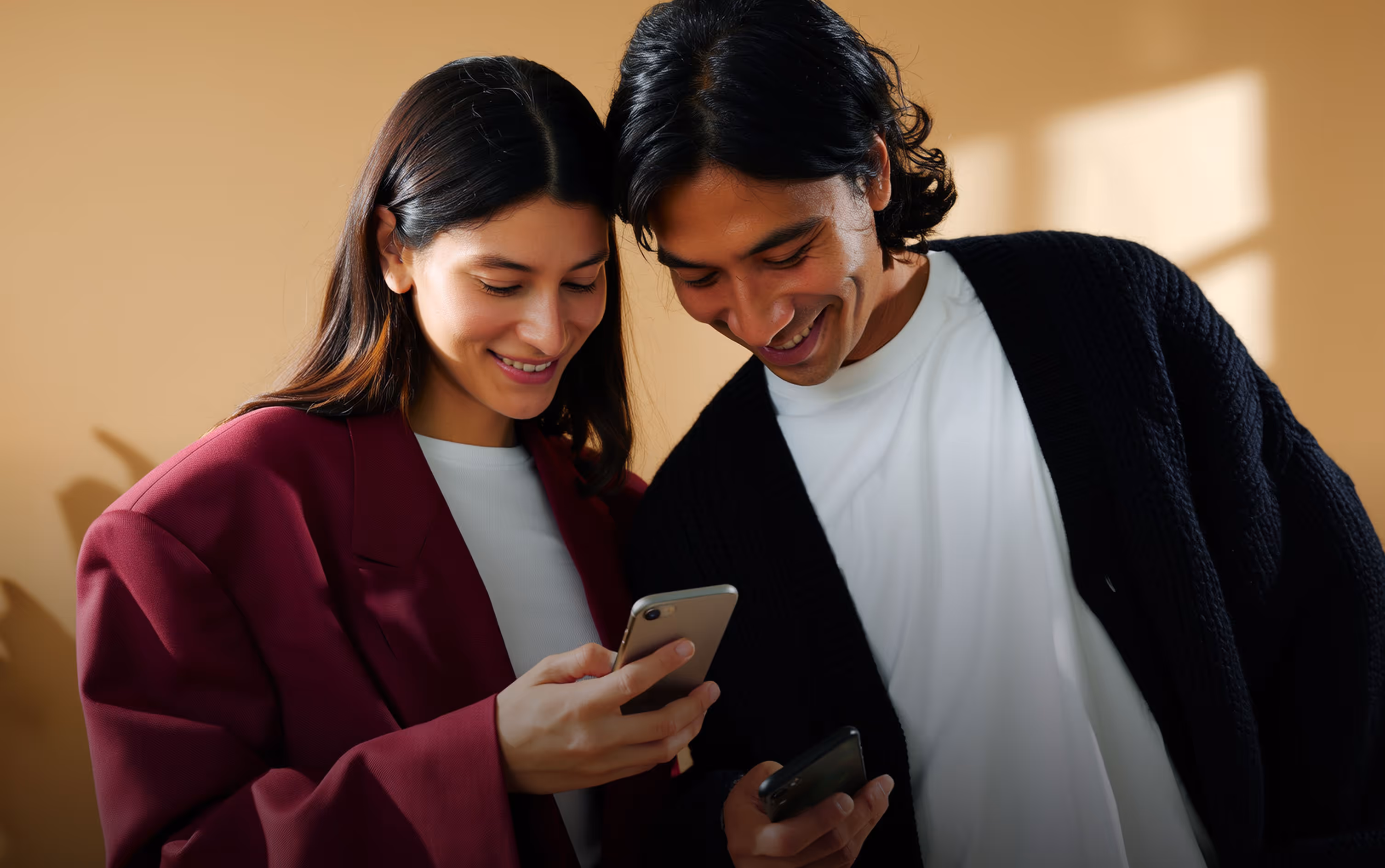 Smiling young woman and man looking at their smartphones together against a beige background.