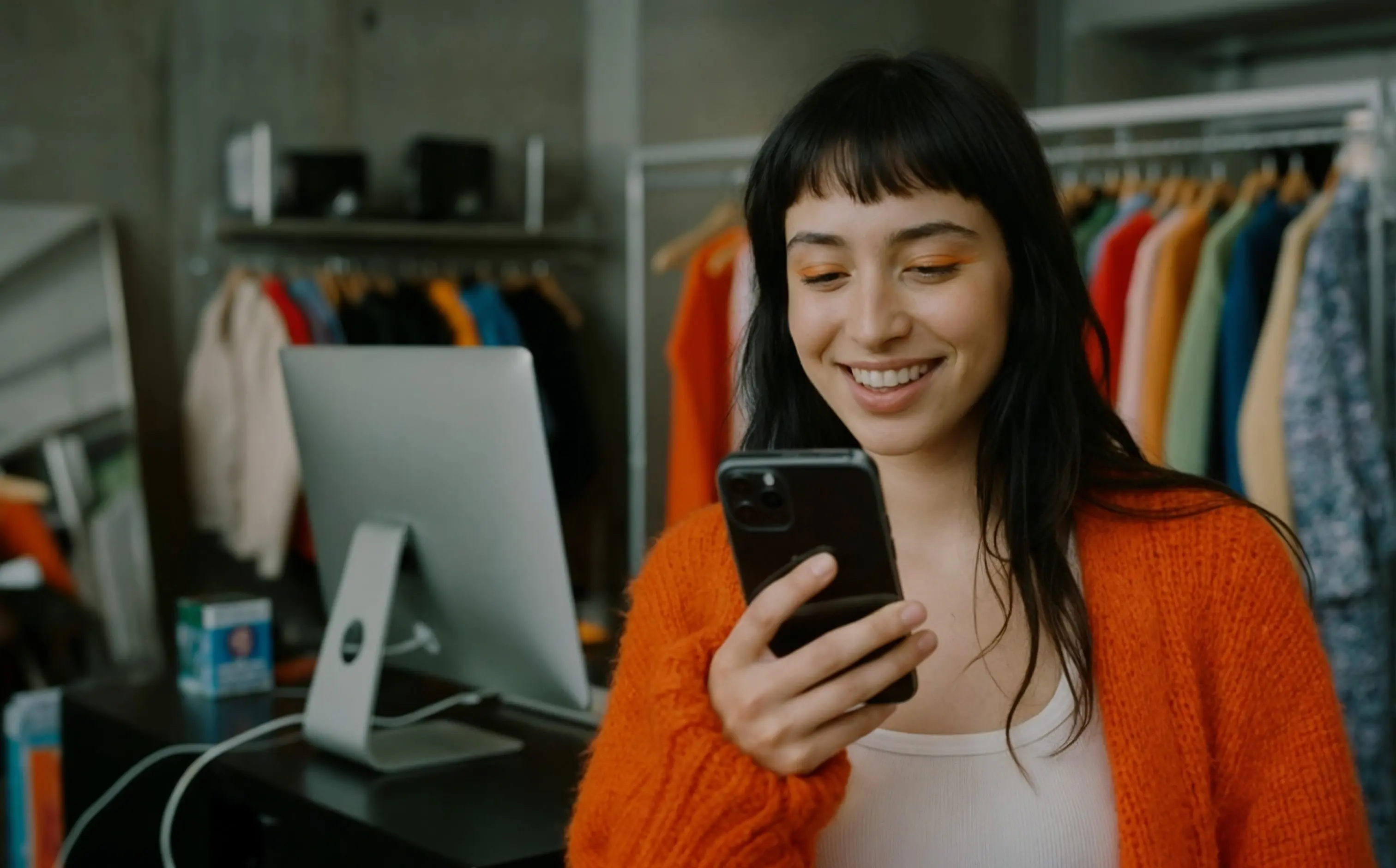 Young woman in orange sweater smiling while looking at her smartphone in a clothing store.