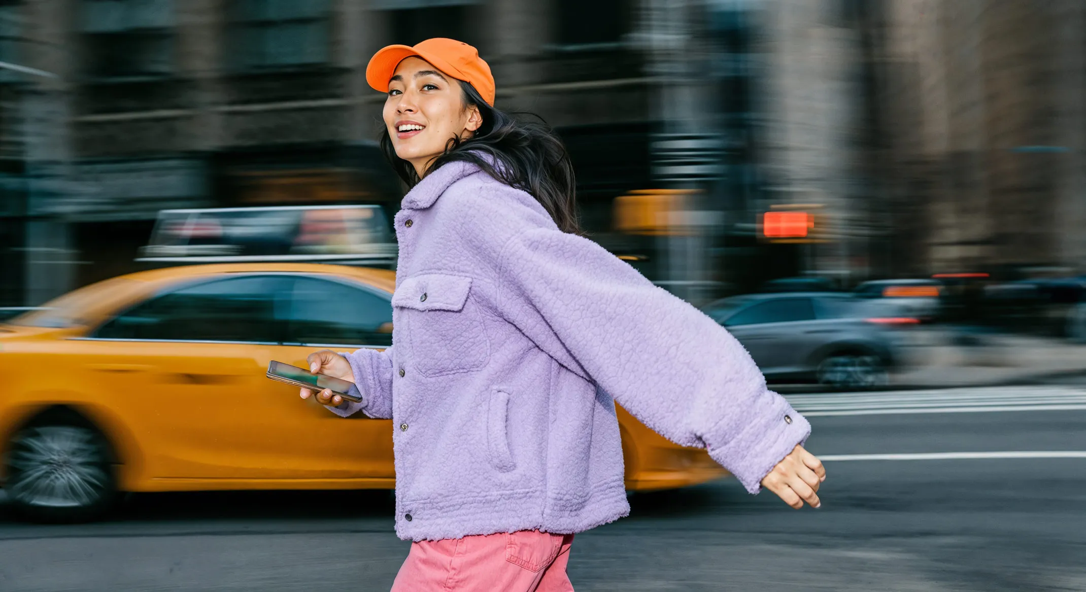 Woman in purple jacket and orange cap walking past a yellow taxi on a city street holding a smartphone.