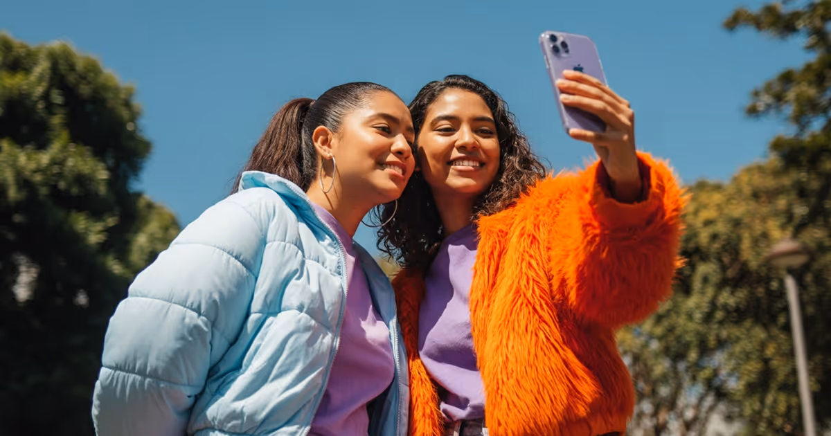 Two young women smiling and taking a selfie outdoors on a sunny day, one wearing a light blue jacket and the other an orange fuzzy coat.