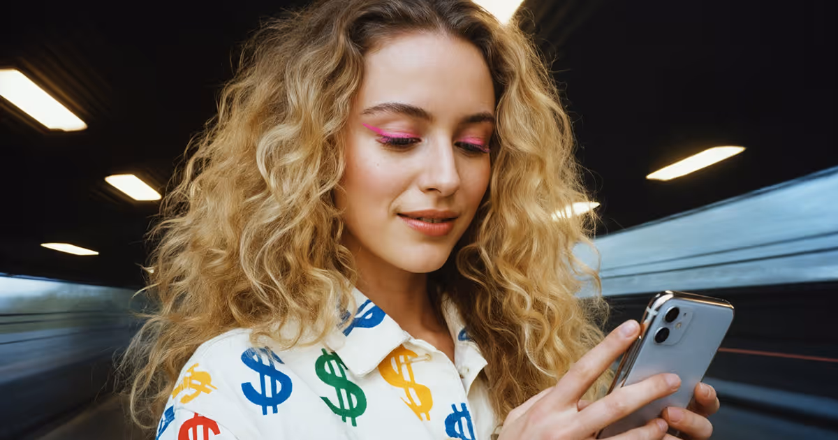 Woman with curly blonde hair wearing a white shirt with colorful dollar signs, looking at a smartphone in her hands.