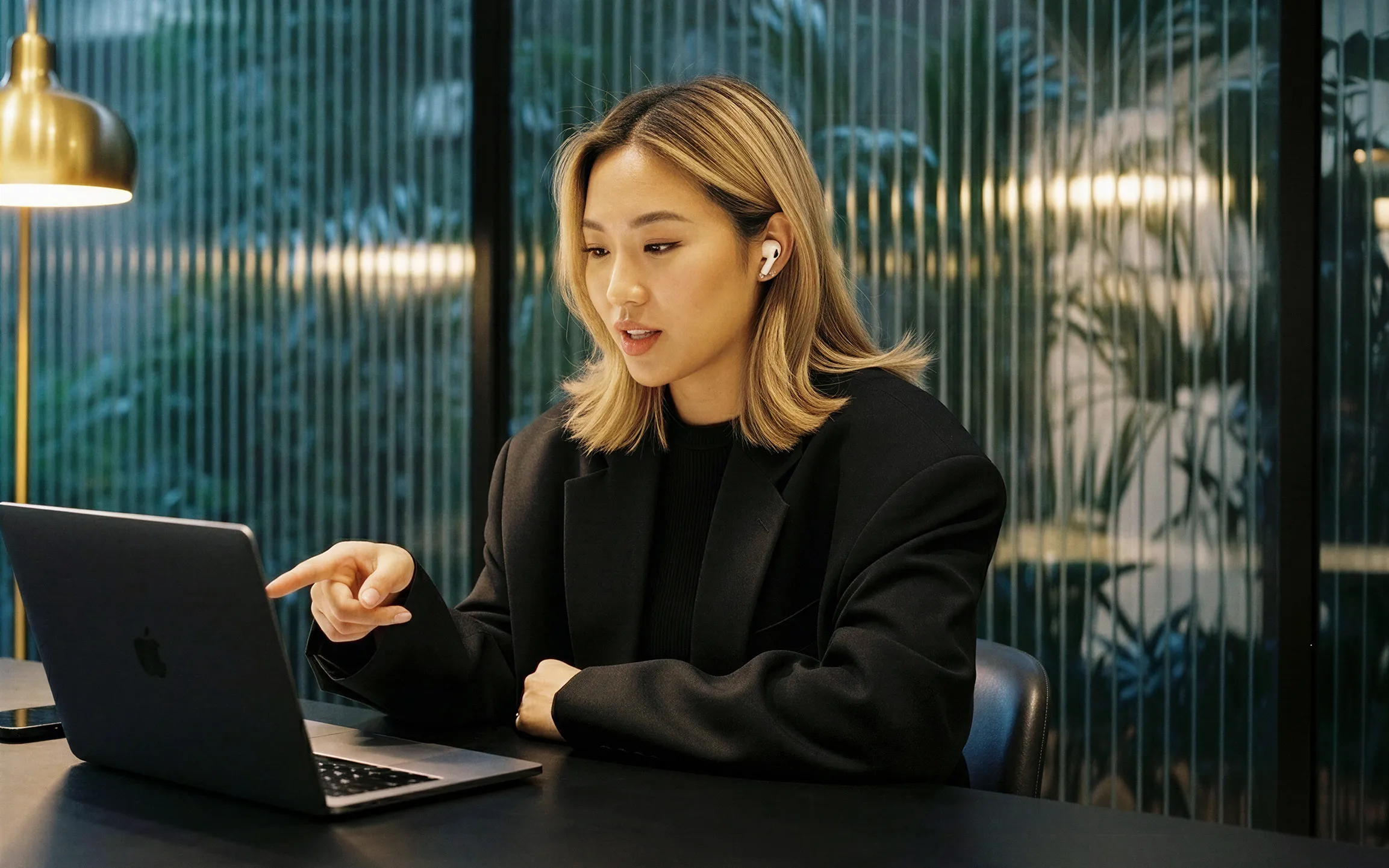 Woman wearing earbuds pointing at a laptop screen while sitting at a table indoors.