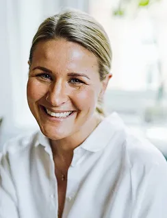 Smiling woman with blonde hair pulled back wearing a white shirt in a bright indoor setting.