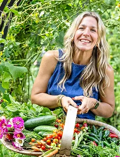 Smiling woman holding basket full of fresh vegetables and flowers outdoors.