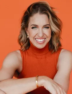 Smiling woman with shoulder-length wavy hair wearing an orange sleeveless top against an orange background.