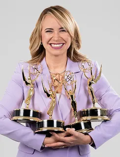 Smiling woman in lavender blazer holding four Emmy awards against a light gray background.