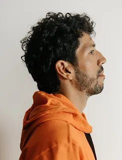 Profile of a man with curly hair and beard wearing an orange hoodie against a neutral background.
