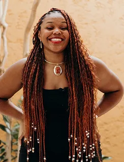 Smiling woman with long braids wearing a black dress and a pendant necklace against a beige wall.
