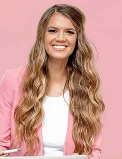Smiling woman with long wavy hair wearing a pink blazer and white top against pink background.