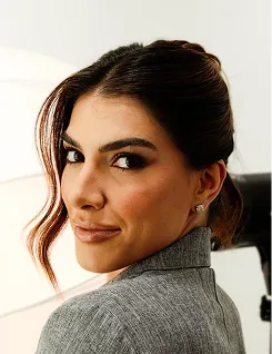 Woman with dark hair and makeup wearing a gray top, looking over her shoulder indoors.