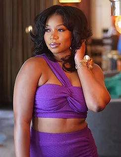 Woman in a purple sleeveless crop top and skirt posing indoors with hand near her neck.