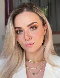 Close-up of a woman with long blonde hair, wearing makeup and layered gold necklaces.