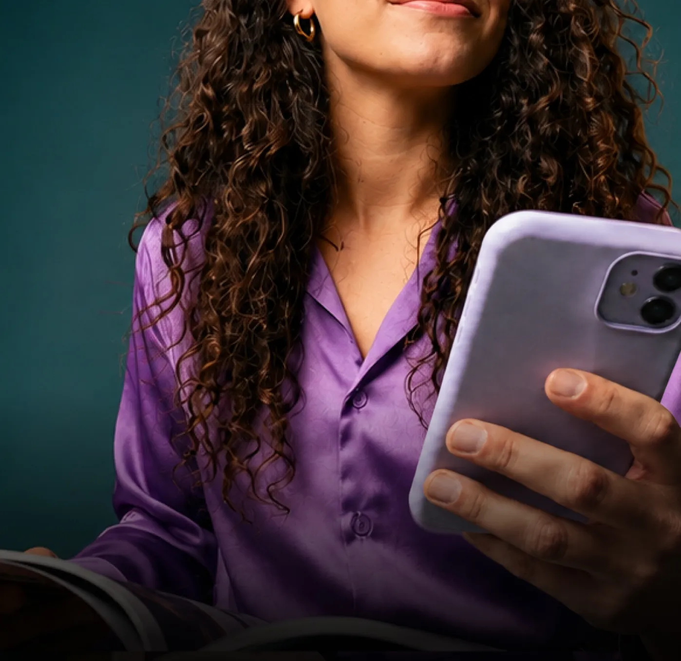 Woman with curly hair in a purple shirt holding a phone with a lavender case and smiling slightly.