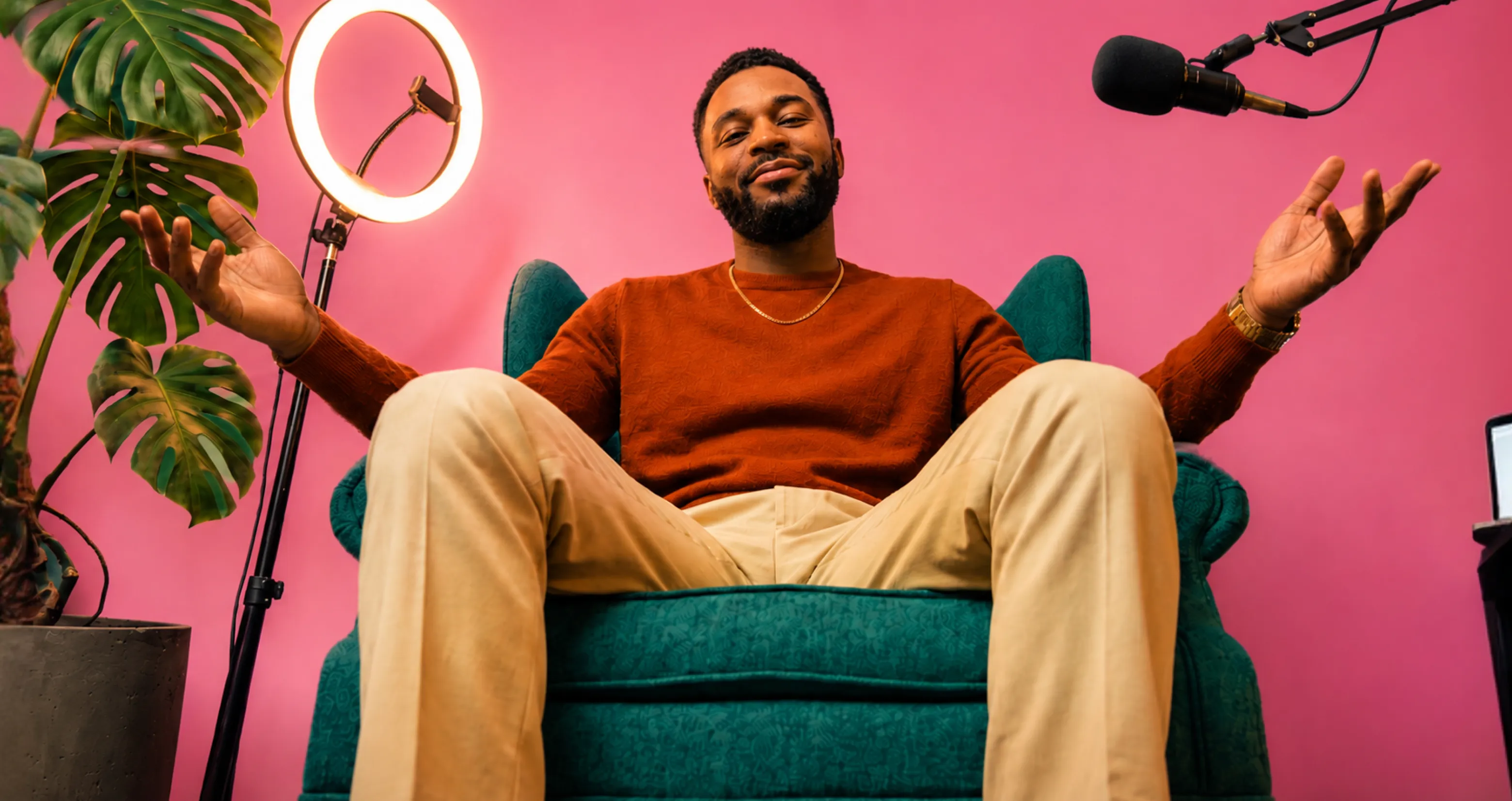 Man in orange sweater sitting on teal chair with open arms, pink background, ring light, and microphone.