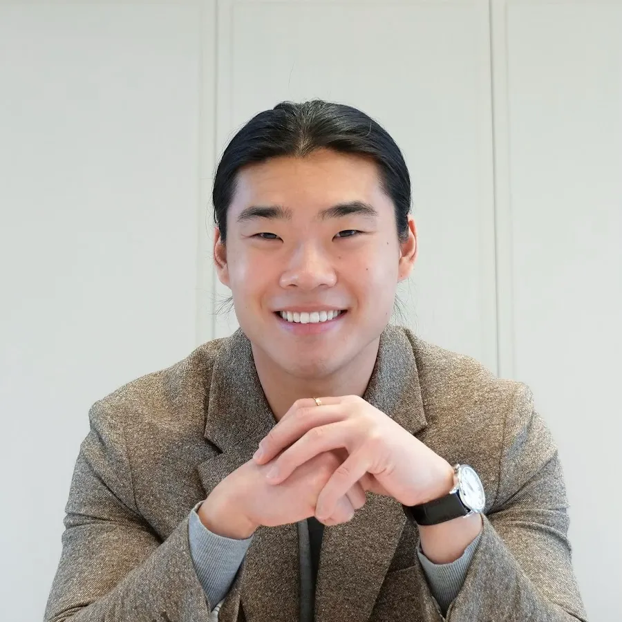 Smiling man with dark hair in a brown blazer and watch, hands clasped in front of him.